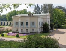 Kleines klassizistisches Gebäude mit weißer Fassade und blauen Fensterläden, umgeben von gepflegtem Garten mit Blumenbeeten und Rasenflächen; im Hintergrund sind Bäume und eine Pergola zu sehen.