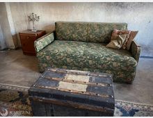 Rustikaler Innenraum mit rau verputzten Wänden und Betonboden: Im Mittelpunkt steht ein grünes, gemustertes Sofa mit Kissen, davor eine große schwarze Truhe mit Metallbeschlägen. Links daneben steht ein kleiner Holz-Nachttisch mit silbernem Kerzenleuchter, teils auf einem gemusterten Teppich.