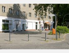 Wohngebäude mit mehreren Fenstern und einem Ladengeschäft im Erdgeschoss, das teilweise mit einer orangefarbenen Markise bedeckt ist. Im Vordergrund ein gepflasterter Gehweg mit Pollern und ein orangefarbener Papierkorb; rechts ist ein Straßenschild mit der Aufschrift „Brusendorfer Str.“ sichtbar.