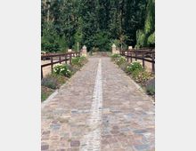 Steinweg mit Pflastersteinen, in der Mitte ein heller Streifen aus kleineren Steinen. Der Weg ist von blühenden Blumen und niedrigen Holzgeländern gesäumt. Im Hintergrund dichter Wald und vereinzelte Backsteinpfeiler.