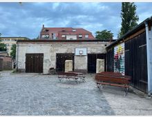 Gepflasterter Hof mit einfacher, leicht verfallener Backsteinfassade, mehreren dunklen Holztoren und einem Basketballkorb an der Wand. Im Vordergrund stehen Holzsitzbänke und Tische, daneben hängt links eine Lichterkette, rechts ein Garagentor mit bunter Graffiti-Bemalung.