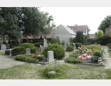 Ein friedlicher Dorffriedhof mit Grabsteinen, gepflegten Gräbern und bunten Blumen. Im Hintergrund stehen Häuser mit roten Ziegeldächern, umgeben von Bäumen und Büschen.