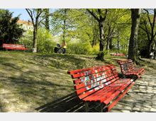 Ein Park mit mehreren roten Bänken, teilweise mit Graffiti bemalt, auf einem mit Steinplatten und Erde bedeckten Untergrund. Im Hintergrund sind Bäume mit grünem Laub, ein Spaziergänger mit Kinderwagen und teilweise sichtbare Gebäude zu sehen.