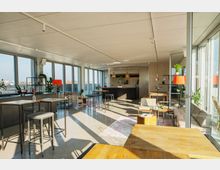 Heller, moderner Multifunktionsraum mit großen Fensterfronten und Blick auf die Stadt. Der Raum ist möbliert mit hohen Tischen, Sitzmöglichkeiten, Regalen mit Pflanzen und einer Bar im Hintergrund, beleuchtet durch natürliches Tageslicht.