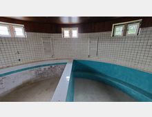 Innenansicht eines leerstehenden, ovalen Pools mit türkisfarbenen Fliesen und einer umlaufenden weißen Fliesenumrandung. Die Wände sind mit weißen Kacheln verkleidet, es gibt mehrere kleine Fenster und zwei Heizkörper an den Wänden.