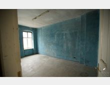 Leerstehender Raum mit bläulich gestrichenen Wänden, einem großen Fenster mit weißem Rahmen und Betonboden. Die Decke ist unverkleidet und weist eine einfache, lange Leuchtstofflampe auf.