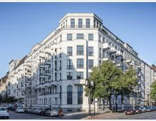 Mehrstöckiges Eckwohngebäude mit heller, klassisch gestalteter Fassade, zahlreichen Fenstern und Balkonen. Vor dem Gebäude stehen Bäume und geparkte Autos, die umliegende Straße ist von Wohngebäuden gesäumt.