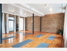 Ein heller Yoga- oder Übungsraum mit Holzfußboden, freiliegenden Ziegelwänden und Metallstützen. Blaue Yogamatten sind gleichmäßig auf dem Boden verteilt, und große Fenster mit weißen Vorhängen lassen Tageslicht herein.