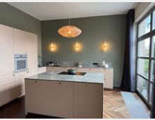 Modern kitchen with pale pink flat-panel cabinets and a large marble-look island featuring a black cooktop, set against a green wall with three wall sconces and a woven pendant light. Floor-to-ceiling windows with dark curtains bring in daylight above a wood herringbone floor and long floor vent.
