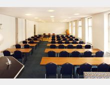 Ein heller Seminarraum mit mehreren Reihen von Holztischen und blauen Stühlen, große Fenster auf der rechten Seite bringen Tageslicht herein, und ein Rednerpult steht vorne im Raum. Die Wände sind weiß, der Boden ist mit Teppich ausgelegt.