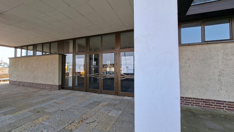 Eingangsbereich eines modernen Gebäudes mit überdachtem Vorplatz, breiten Steinplatten und einer großen weißen Stützsaule im Vordergrund. Dahinter befinden sich mehrere braune Glastüren mit seitlichen Fensterfronten und verputzten Außenwänden mit Sockel aus rotem Ziegelstein.