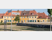 Straßenszene in einem Wohnviertel mit einem Gebäudeensemble aus gelben Häusern, roten Ziegeldächern und grünen Fensterläden. Im Vordergrund stehen Straßenschilder mit den Namen "Hermann-Boian-Str." und "Albert-Fellert-Str.", umgeben von einer Rasenfläche und einer niedrigen Steinmauer.