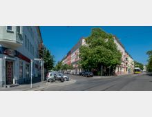 Straßenszene in einer Wohngegend mit mehrstöckigen, farbigen Wohnhäusern, die von Bäumen gesäumt sind. Im Vordergrund stehen parkende Autos und Motorräder, rechts ist eine Straßenbahn auf den Gleisen zu sehen.