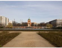 Blick über einen städtischen Parkteich mit einem geraden Weg im Vordergrund; gegenüber steht ein markanter Backsteinturm mit grüner Kuppel und großer Rundöffnung. Links und rechts rahmen mehrgeschossige Wohn- und Bürogebäude sowie kahle Bäume das Ufer.