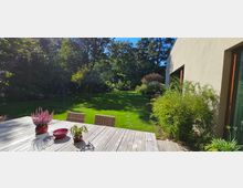 Garten mit einer großen Rasenfläche, umgeben von dichtem Grün und hohen Bäumen. Im Vordergrund steht ein Holztisch mit Dekorationen, daneben ein modernes Gebäude mit großen Fenstern und Pflanzen am Rand.