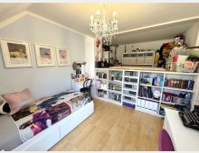 Helles Dachzimmer als Schlafzimmer mit Holzboden, weißem Bett und Schreibtisch sowie einem Kristall-Kronleuchter an der Decke. Entlang der Wand steht ein großes weißes Regalsystem mit Büchern, Boxen und Spielzeug, darüber ein erhöhtes Podest mit weiteren Aufbewahrungsmöbeln.