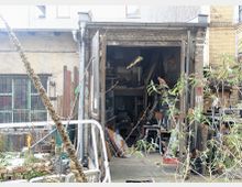 Hof mit einem offenen Werkstattschuppen, umgeben von alten Gebäuden aus Ziegelmauerwerk. Im Inneren der Werkstatt sind Werkzeuge, Regale und Materialien erkennbar, während Pflanzen und Gegenstände den Hof teils verdecken.