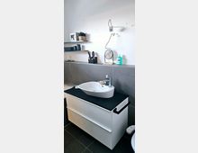 Moderne Badezimmer-Ecke mit einem weißen Waschtisch, darauf ein ovales Waschbecken und Pflegeprodukte. Im Hintergrund sind graue Fliesen, eine weiße Wand mit Regalen sowie ein runder, kleiner Spiegel zu sehen.