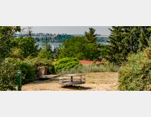 Ein Spielplatz mit einer alten Karussellstruktur in einer sandigen Umgebung, umgeben von dichten Büschen und Bäumen. Im Hintergrund sind ein See, Wälder und eine Hügellandschaft mit Windrädern und Gebäuden sichtbar.