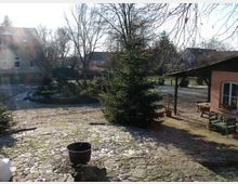 Ein gepflasterter Hof mit einem großen Nadelbaum in der Mitte, umgeben von kahlen Bäumen und Gebäuden mit Ziegeldächern im Hintergrund. Rechts im Bild befindet sich eine überdachte Sitzecke neben einem kleinen, orange gestrichenen Gebäude.