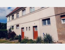 Zweistöckiges Reihenhaus mit cremefarbener Fassade, roten Eingangstüren und dekorativen weiß gerahmten Fenstern, umgeben von einem kleinen Vorgarten mit Pflanzen und einem Gehweg.