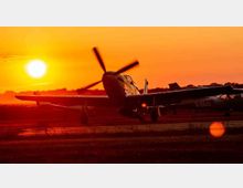 Ein Propellerflugzeug steht auf einem Flugfeld bei Sonnenuntergang, mit einer intensiv orangefarbenen Himmelsszene und einer tiefstehenden Sonne im Hintergrund. Silhouetten von weiteren Flugzeugen und Bäumen sind am Horizont zu erkennen.