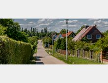 Ein ruhiger, ländlicher Weg mit Laternen, gesäumt von Büschen und Zäunen, führt an Einfamilienhäusern mit roten und grauen Schrägdächern vorbei. Im Hintergrund ist eine Baumreihe sowie ein teilweise bewölkter Himmel zu sehen.