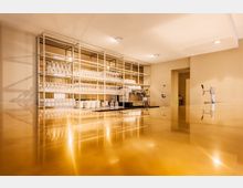 Modernes Café mit einem glänzenden Tresen aus goldfarbenem Material. Im Hintergrund stehen Regale mit gestapelten Gläsern, Geschirr und einer Kaffeemaschine.