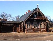 Holzhaus im Blockhausstil mit verzierten Giebelkanten, einer überdachten Terrasse und Fenstern mit Fensterläden. Das Gebäude steht an einer Straße, im Hintergrund sind kahle Bäume sichtbar.
