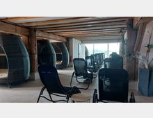 Raum mit Entspannungsliegen und runden geflochtenen Sitzkabinen aus Rattan, Holzträgern an der Decke und einer großen Fensterfront, die Tageslicht hereinlässt und eine Aussicht auf die Landschaft bietet.