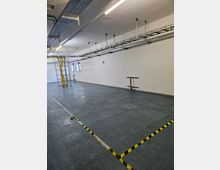 Industriehalle mit grauem Bodenbelag, weißen Wänden und einer Decke mit Kabeltrassen und Beleuchtung. Eine Leiter steht neben gelben Kabeln, und Warnmarkierungen trennen Bereiche des Bodens.