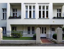 Fassadenansicht eines weißen Wohnhauses im Stil der Gründerzeit mit dekorativen Stuckelementen über den Fenstern, einem kleinen Vorgarten, einer Metallzaunabgrenzung und einem gepflasterten Eingangsweg. Das Haus verfügt über hohe, schmale Fenster und einen überdachten Eingangsbereich.