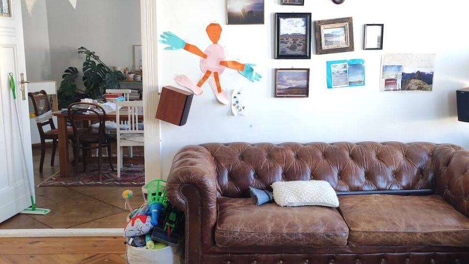 A cozy living room with a vintage brown leather sofa against a white wall adorned with framed pictures and colorful artwork. A wicker coffee table holds books and items, and an open doorway reveals a dining area with wooden furniture and indoor plants.