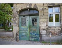 Eine alte, verwitterte Hausfassade mit einer grünen, doppelten Holztür, verziert mit Metallgittern und Graffiti. Rechts neben der Tür befindet sich ein Fenster, links führt der Blick auf einen begrünten Bereich mit Bäumen und einer Sitzgruppe.