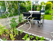 Terrasse aus Holz mit angrenzendem Hochbeet, bepflanzt mit Gemüse und Kräutern. Im Hintergrund ein Metallzaun, Rasenflächen und ein Wohnhaus.