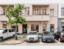 Straßenansicht eines hell verputzten Mehrfamilienhauses mit mehreren Fenstern und Balkonen sowie Ladenfronten im Erdgeschoss. Davor stehen ein Straßenbaum, eine Laterne und mehrere parkende Autos entlang des Bordsteins.