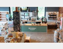 Innenansicht einer modernen Markthalle mit einem Cafébereich. Im Vordergrund sind Waren wie Eier, Getränke und Honig auf Holzkisten ausgestellt, während im Hintergrund eine grüne Theke mit Kaffeemaschine, Preistafel und Regalen mit Produkten zu sehen ist; große Fenster lassen Tageslicht herein.