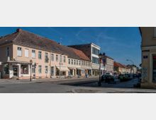 Straßenszene in einer Kleinstadt mit einer Reihe von historischen Wohn- und Geschäftsgebäuden, teils mit Schaufenstern und Markisen. Auf der gepflasterten Straße und den Bürgersteigen stehen geparkte Autos, Straßenlaternen und vereinzelt Pflanzenkübel.