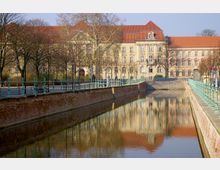 Historisches Gebäude mit rotem Ziegeldach an einem Kanal, umgeben von kahlen Bäumen. Der Kanal ist von roten Backsteinmauern eingefasst, und eine Brüstung mit grünen Geländern verläuft entlang des Gehwegs.
