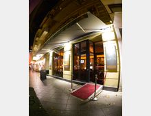 Außenansicht einer Bar oder eines Cafés bei Nacht mit gelblicher Fassade, großen verglasten Türen und Fenstern, einer Markise und einem roten Teppich mit Absperrseilen vor dem Eingang. Die Straße ist gepflastert und von herbstlichem Laub bedeckt.