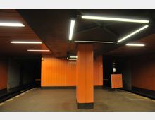 U-Bahn-Station mit orangefarbenen Wand- und Säulenverkleidungen, einer dunklen Bahnsteigkante und geometrischen Deckenleuchten. Die Atmosphäre ist gedämpft durch das geringe Licht und die dunklen Bereiche im Hintergrund.