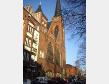 Backsteinkirche im neugotischen Stil mit großen Spitzbogenfenstern und einem hohen Turm mit Uhr vor einem klaren blauen Himmel; links angrenzendes Wohngebäude und geparkte Autos im Vordergrund.