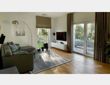 Modernes Wohnzimmer mit grauem Ecksofa, rundem Couchtisch auf Teppich und wandmontiertem Fernseher über einem schwebenden Sideboard. Große Glas-Terrassentüren und ein schmales Fenster mit Vorhängen lassen Tageslicht herein und geben den Blick in einen grünen Garten frei.