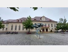 Historisches Gebäude mit Bibliothek, erkennbar durch den Schriftzug „BIBLIOTHEK“ über dem Eingang, mit großen Fenstern und Walmdach. Davor ein gepflasterter Platz mit mehreren geparkten Fahrrädern, einem Baum mit Sitzbereich und einem modernen Glasdach.
