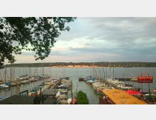 Blick auf einen Jachthafen an einem See mit zahlreichen Segelbooten und Liegeplätzen. Im Vordergrund befinden sich Terrassen mit Sonnenschirmen eines Restaurants, im Hintergrund ist eine bewaldete Uferlandschaft zu sehen.