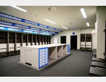 Umkleideraum eines Sportstadions mit offenen Schließfächern an den Wänden, einer großen weißen Sitzbank in der Mitte und blauen Schriftzügen mit Motivationswörtern an den Wandpaneelen. Die Sitzflächen bestehen aus dunklen Polstern, und der Raum ist gut beleuchtet.