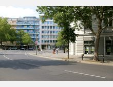 Straßenkreuzung in einer Stadt mit modernen mehrstöckigen Gebäuden, darunter ein blau gestrichenes Wohnhaus mit Balkonen. Der Bereich ist von Bäumen gesäumt, und es sind mehrere Autos, Fußgänger und ein Gehweg sichtbar.
