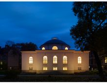 Historisches Gebäude mit weißer Fassade und rundem Kuppeldach, beleuchtet durch große, bogenförmige Fenster in der Dämmerung; umgeben von Bäumen und weiteren Gebäuden im Hintergrund.