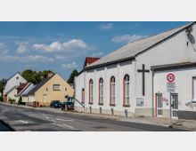 Straßenszene mit einem weißen Gebäude auf der rechten Seite, das eine Fassade mit hohen Rundbogenfenstern und einem Kreuz aufweist, vermutlich eine Kirche oder ein Gemeindesaal. Links daneben stehen Einfamilienhäuser mit Satteldächern, während ein Straßenschild auf eine Geschwindigkeitsbegrenzung von 30 km/h hinweist.