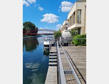 Holzsteg am Ufer eines Flusses mit festgemachtem Boot, daneben ein modernes Gebäude mit Balkonen und Glasfronten; im Hintergrund eine rote Fußgängerbrücke und üppige Vegetation unter blauem Himmel mit wenigen Wolken.